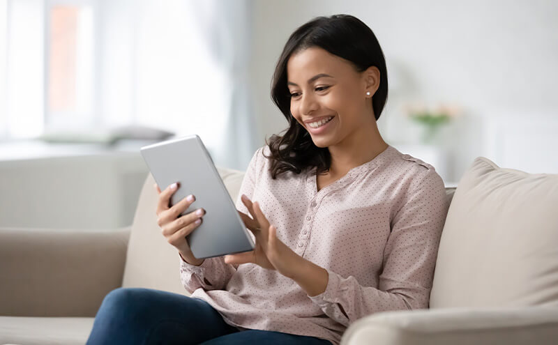 A smiling woman on a couch uses a tablet to make a doctor's appointment.