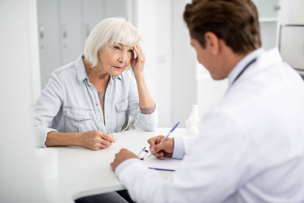 An elderly female patient at a desk speaks with a doctor about vertigo.