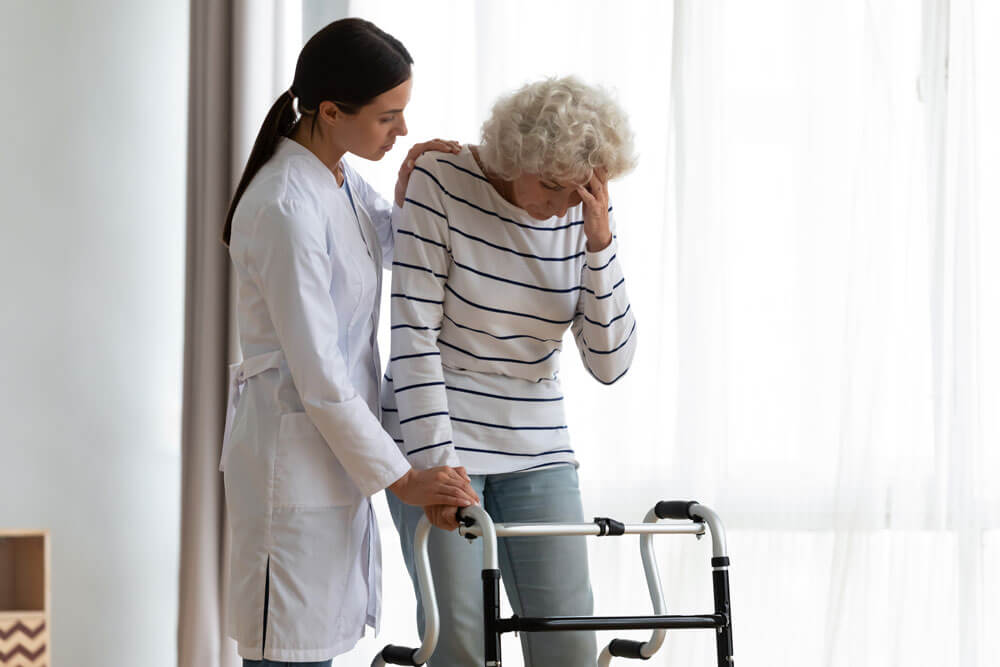 An aide assists an elderly woman with a walker who seems to be experiencing vertigo or dizziness.