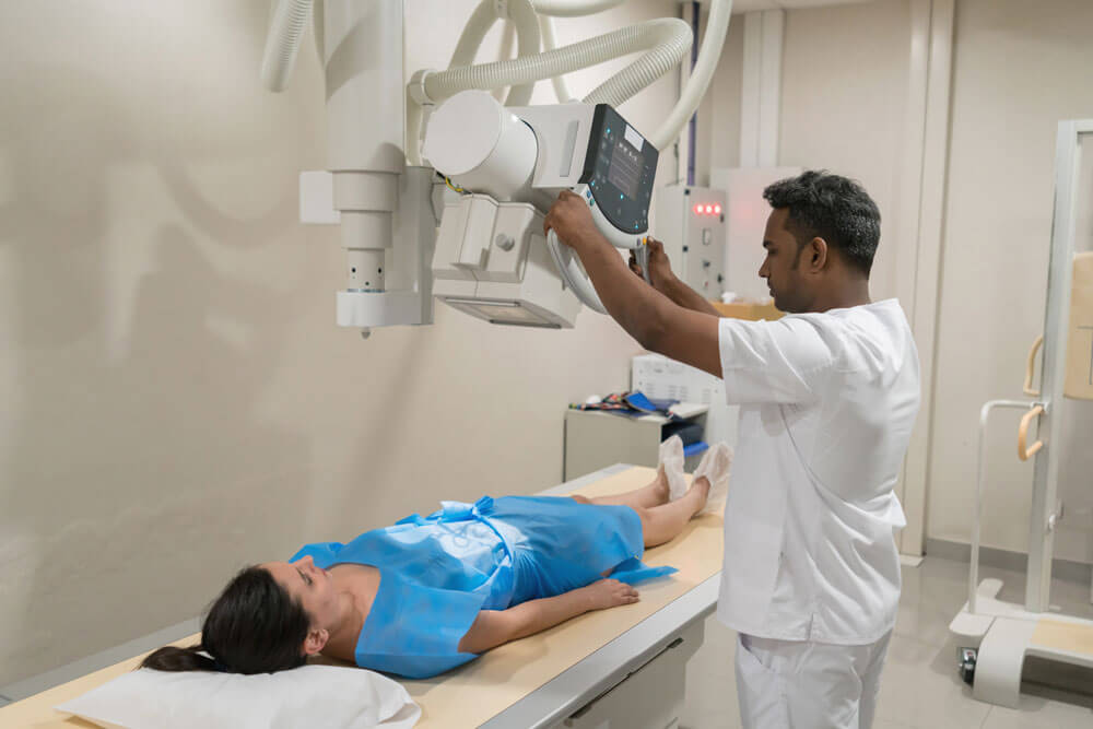 A patient lies on an X-ray table while a radiologist adjusts the imaging equipment.